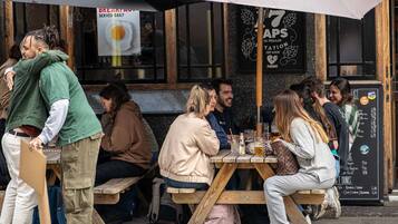 A group of people sitting at a wooden table outside a restaurant with a menu board visible.