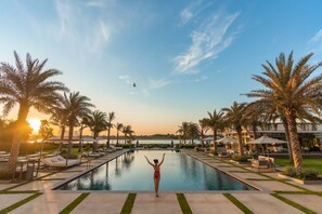 Zona de piscina con sillones de descanso y palmeras, una persona de brazos alzados, y un atardecer al fondo.