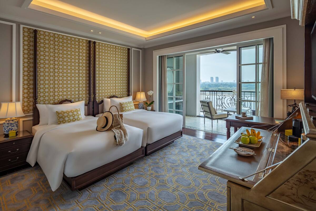 Une chambre d’hôtel avec un grand lit, un bureau agrémenté d’un panier de fruits et un balcon offrant une vue sur la ville.
