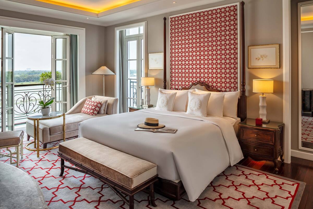 Une chambre spacieuse avec un grand lit, un banc, une table de chevet et une commode. Elle comporte deux fenêtres avec des rideaux, une vue sur la verdure et un mur décoré de motifs.