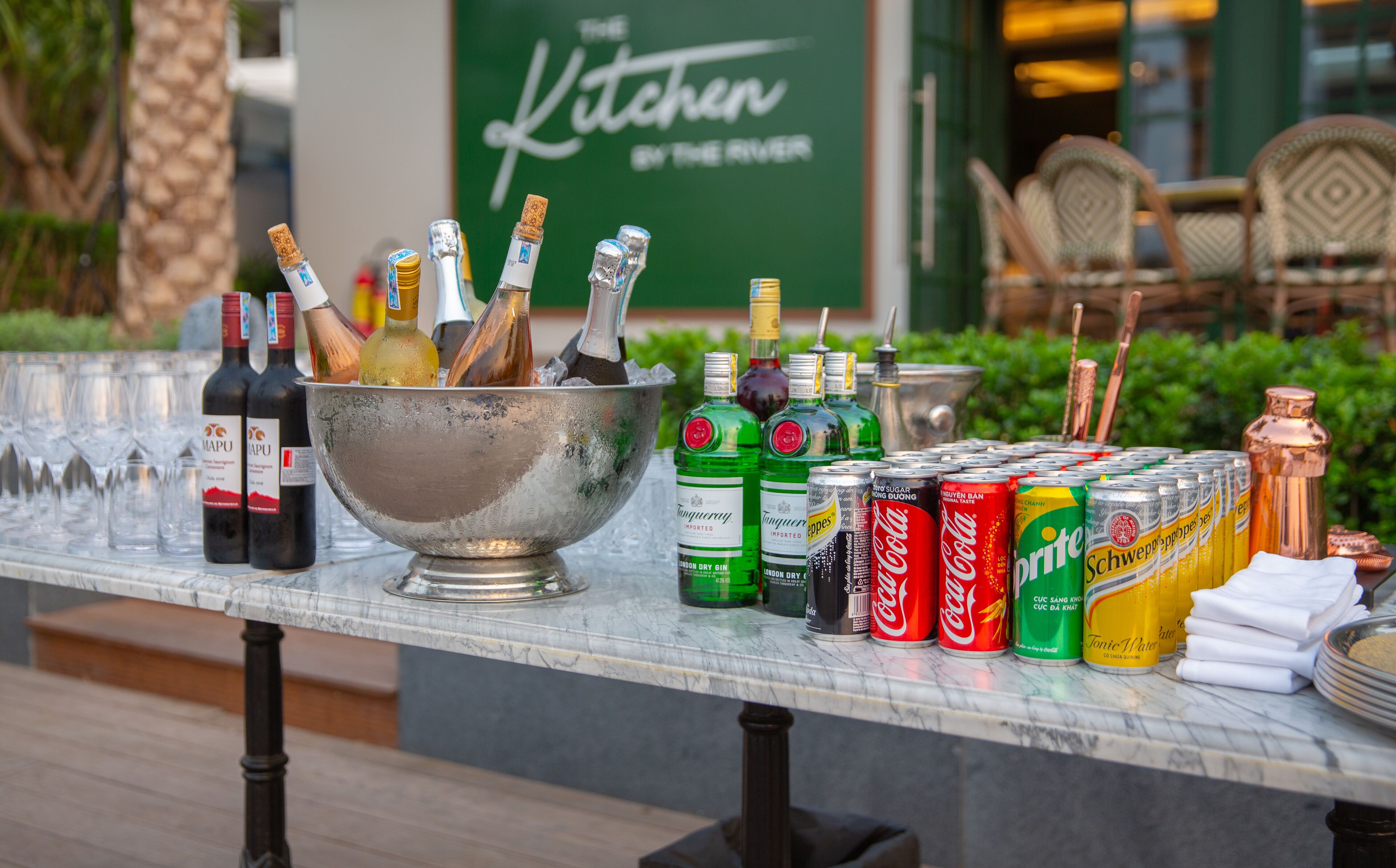 A table with various bottles and cans, including wine, gin, and soda, in an outdoor setting.