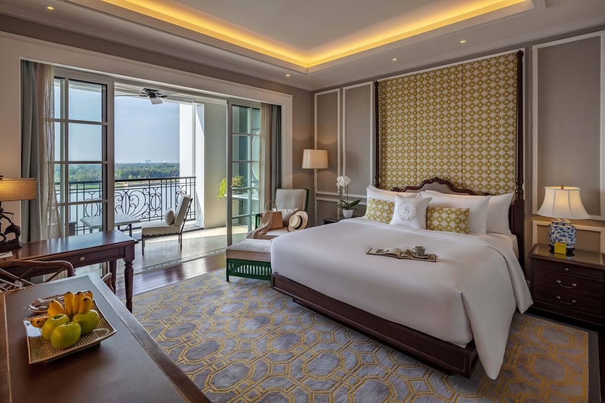 Une chambre d’hôtel avec un grand lit, un bureau agrémenté d’un plateau de fruits, un balcon avec vue et un tapis à motifs.