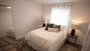 A bedroom with a large bed, two bedside lamps, a window with sheer curtains, and a mirror on the wall.