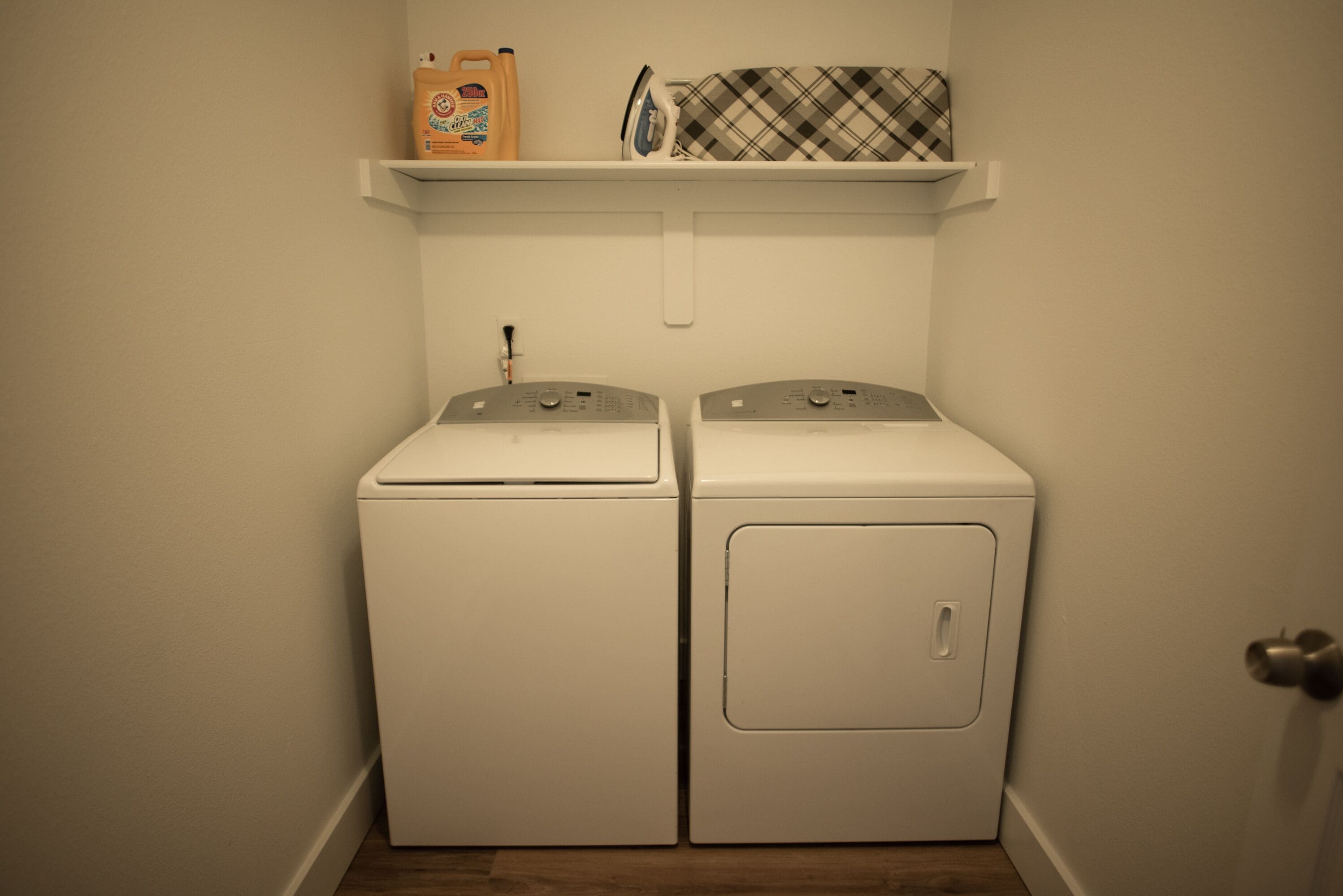 A laundry room with two front-loading washing machines and a shelf above them holding laundry supplies.