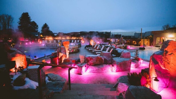 An outdoor heated pool area with seating and rock formations, illuminated by pink lighting.