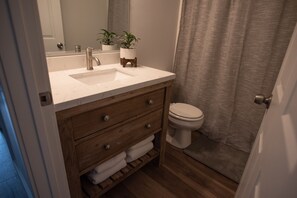Un baño con tocador de madera, un lavabo blanco, dos plantas en macetas, un inodoro y una cortina de ducha.