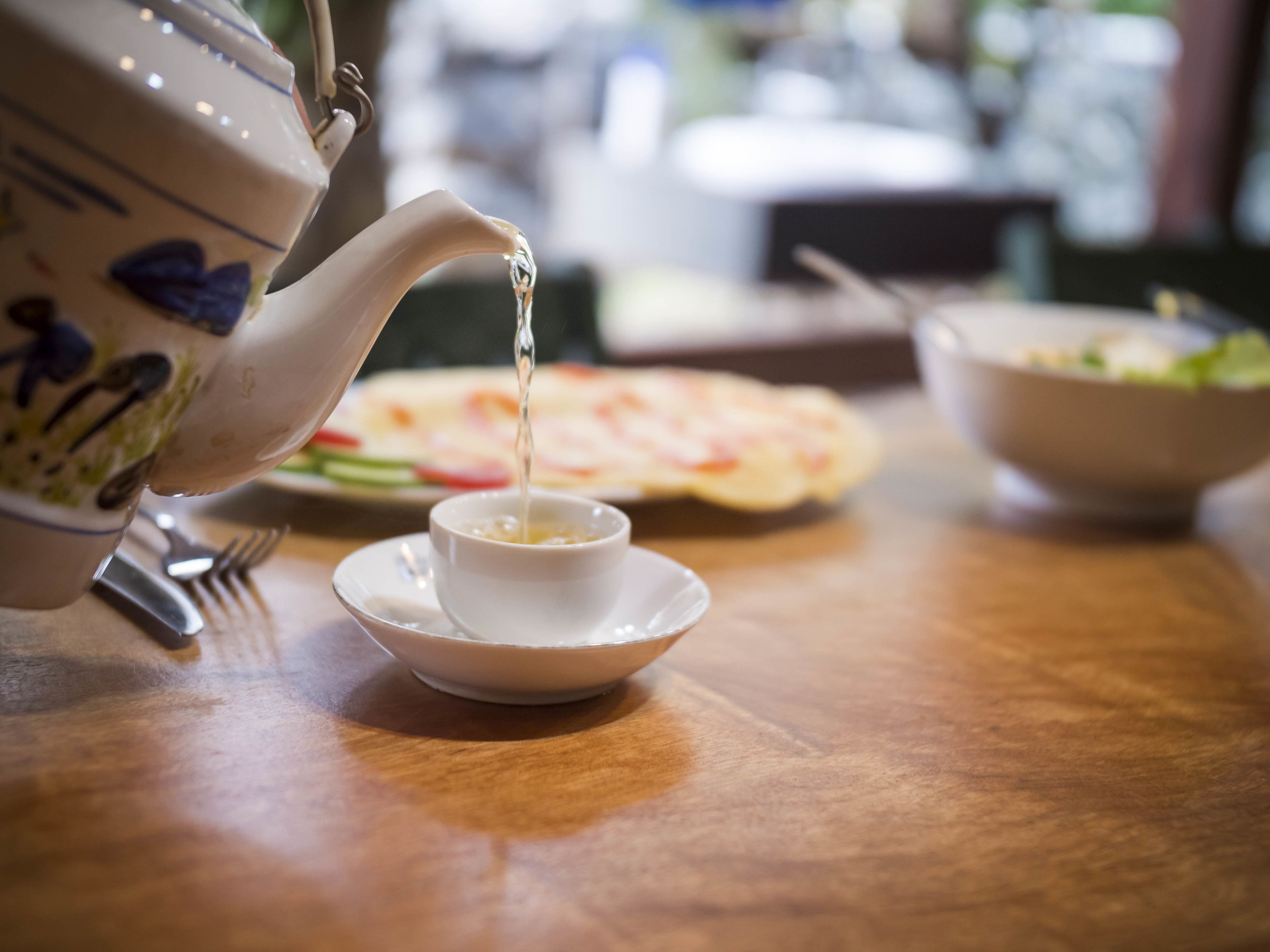 A teapot pouring tea into a cup on a wooden table with a plate of food and a bowl in the background.