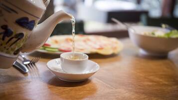 A teapot pouring tea into a cup on a wooden table with a plate of food and a bowl in the background.