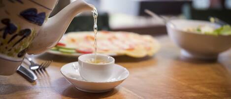 A teapot pouring tea into a cup on a wooden table with a plate of food and a bowl in the background.