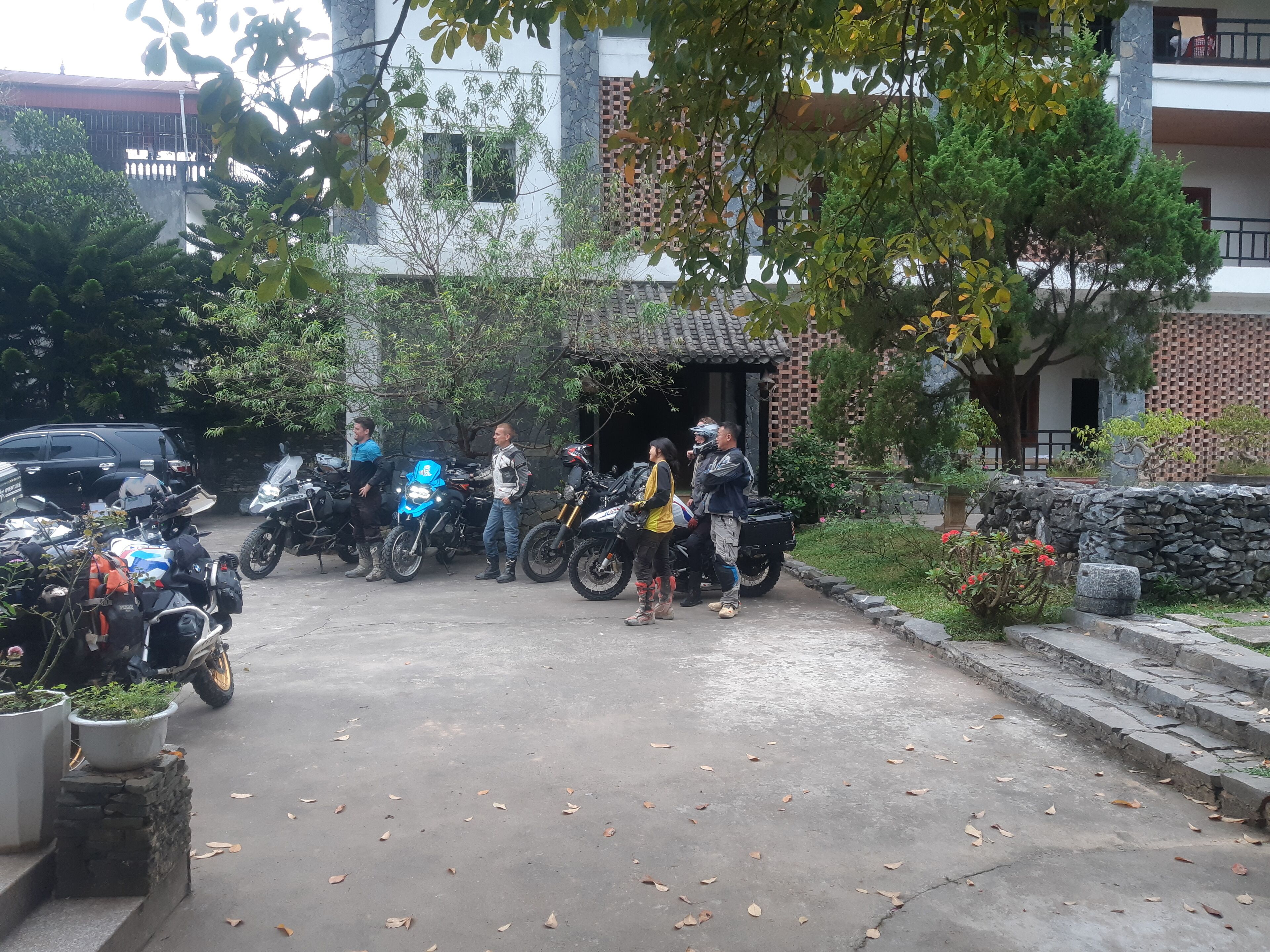 A group of people standing near motorcycles in a courtyard with stone walls and a tiled roof structure.