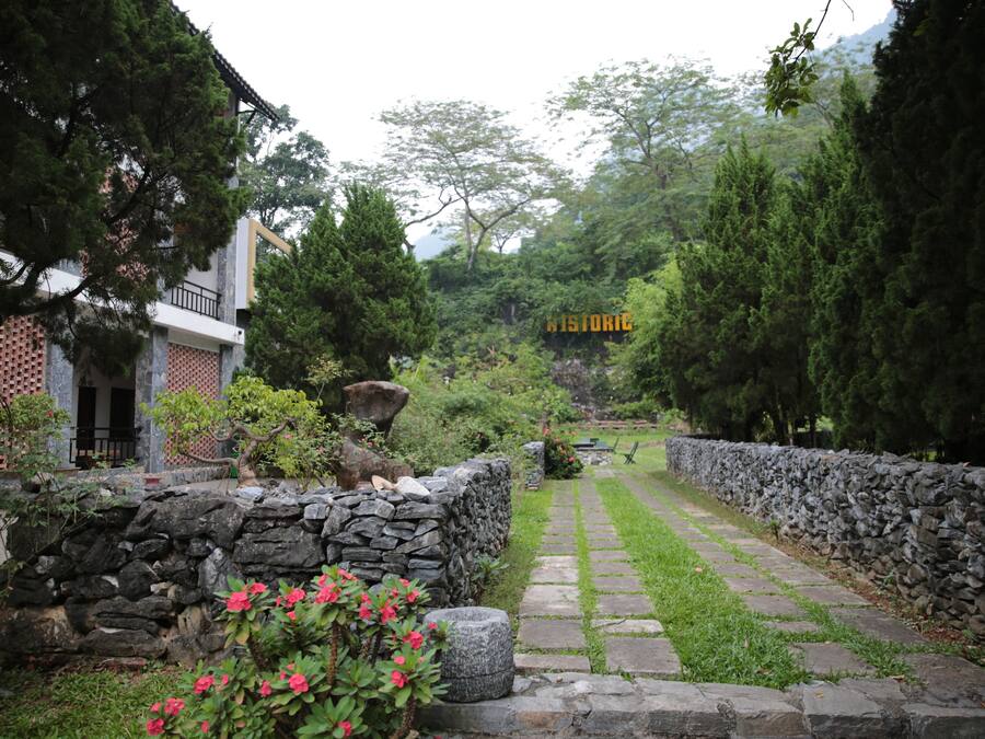 Un chemin de pierre traverse un jardin, avec un mur de pierre d’un côté et un bâtiment doté d’un balcon de l’autre.