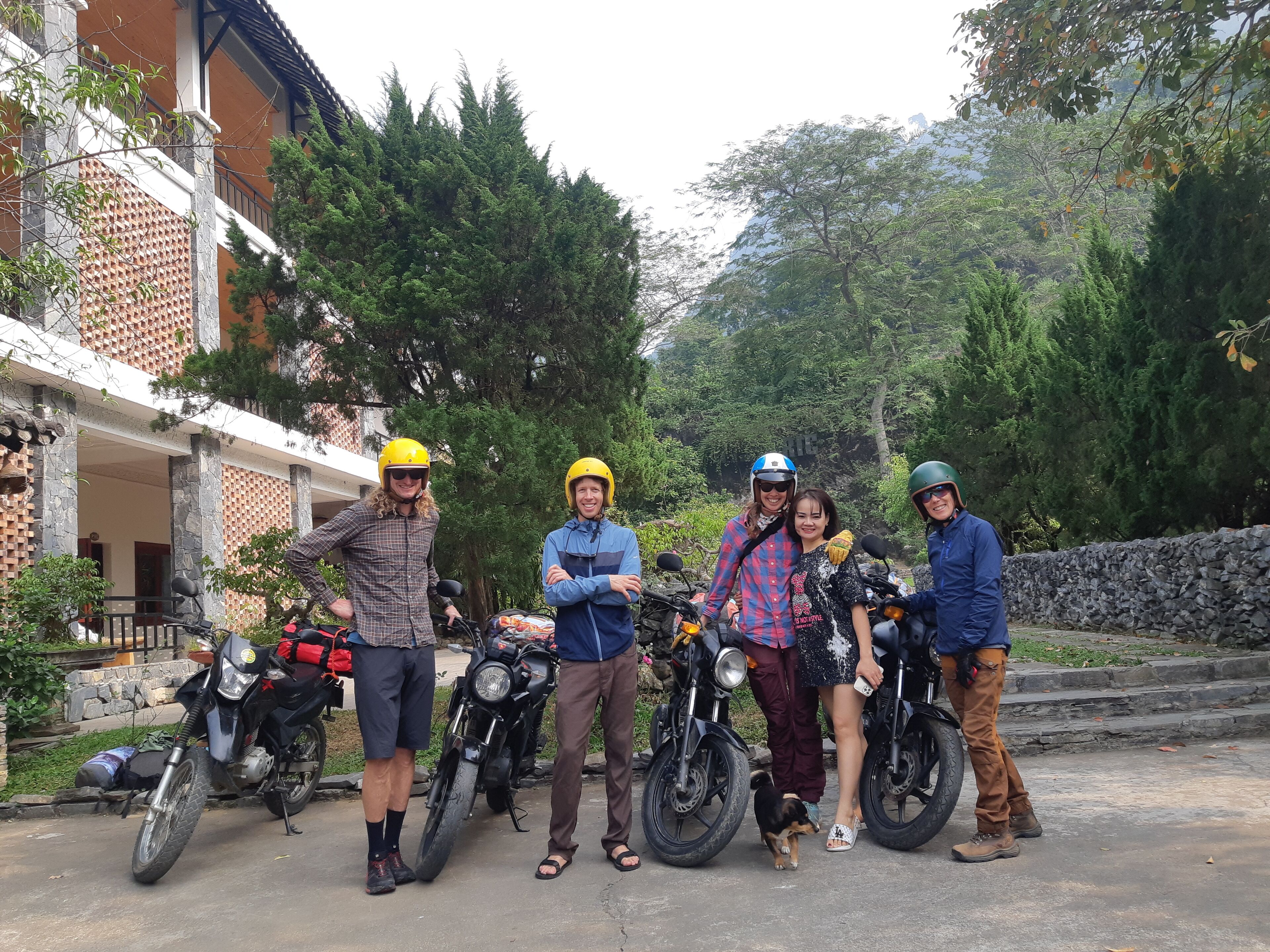 Un gruppo di persone con caschi in piedi accanto a motociclette in un ambiente esterno con alberi e un edificio sullo sfondo.