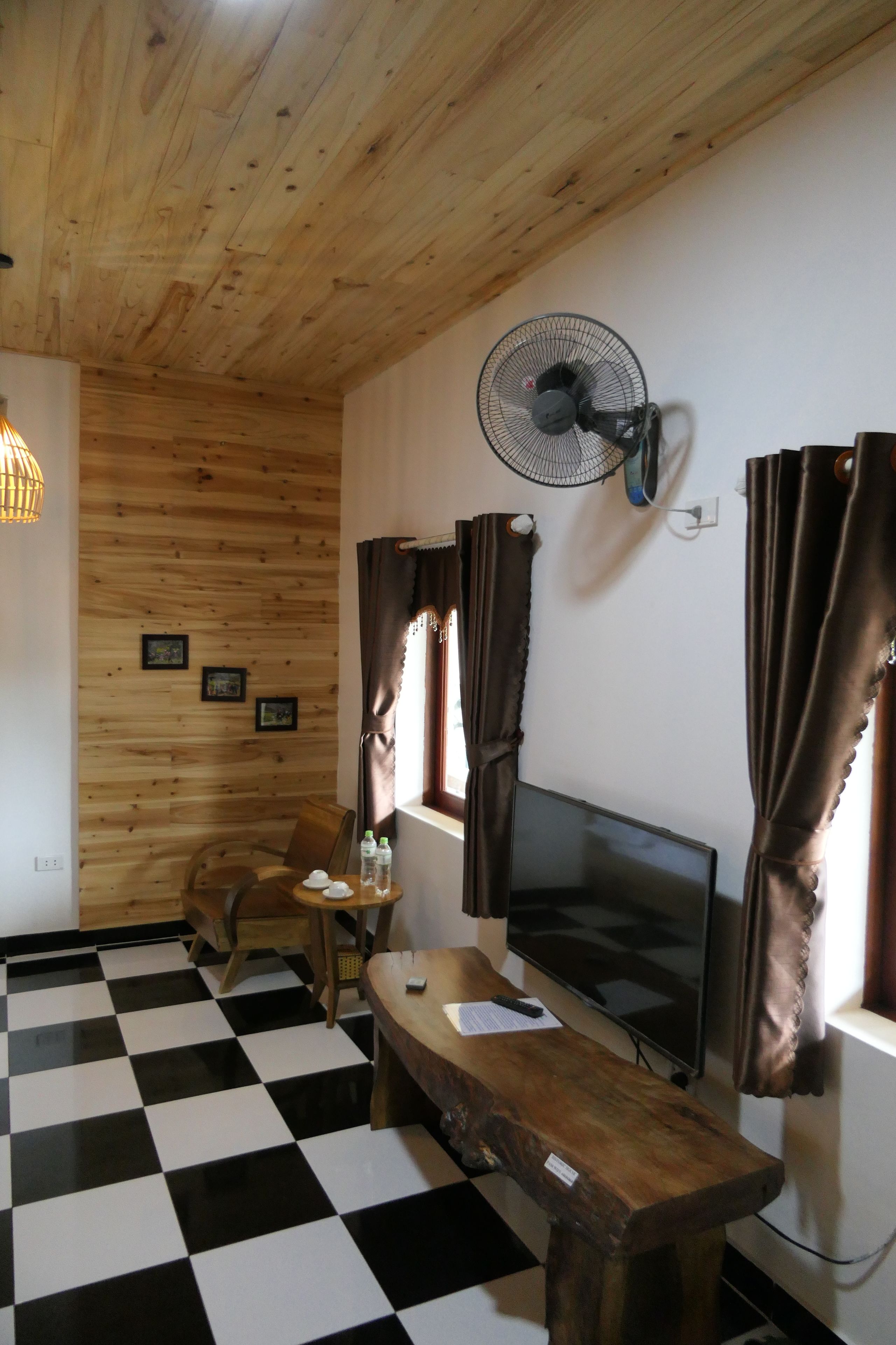 Une pièce avec un plafond en bois, un sol à carreaux noirs et blancs, une table en bois, un téléviseur, un ventilateur et des rideaux.