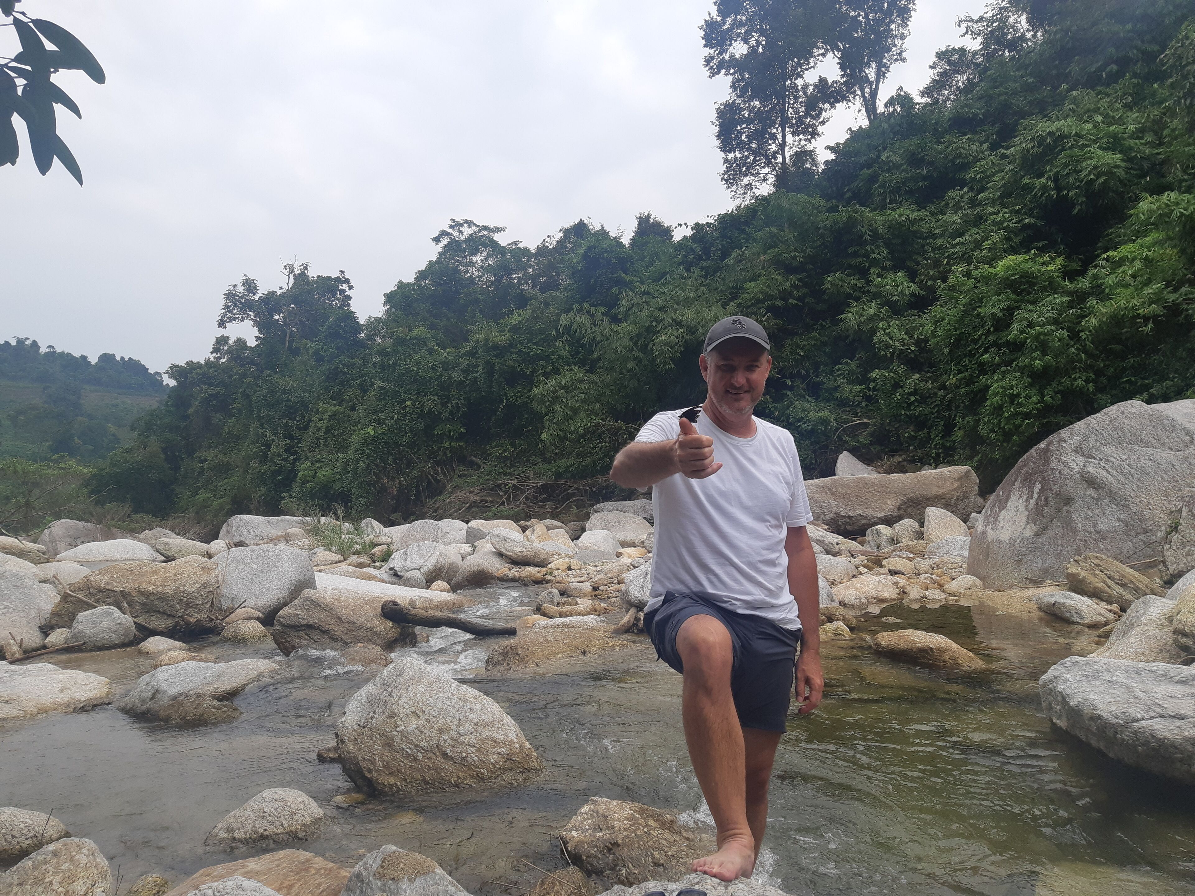 Una persona de pie en un arroyo pedregoso con grandes rocas y abundante vegetación al fondo.