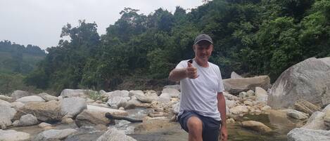 Una persona de pie en un arroyo pedregoso con grandes rocas y abundante vegetación al fondo.