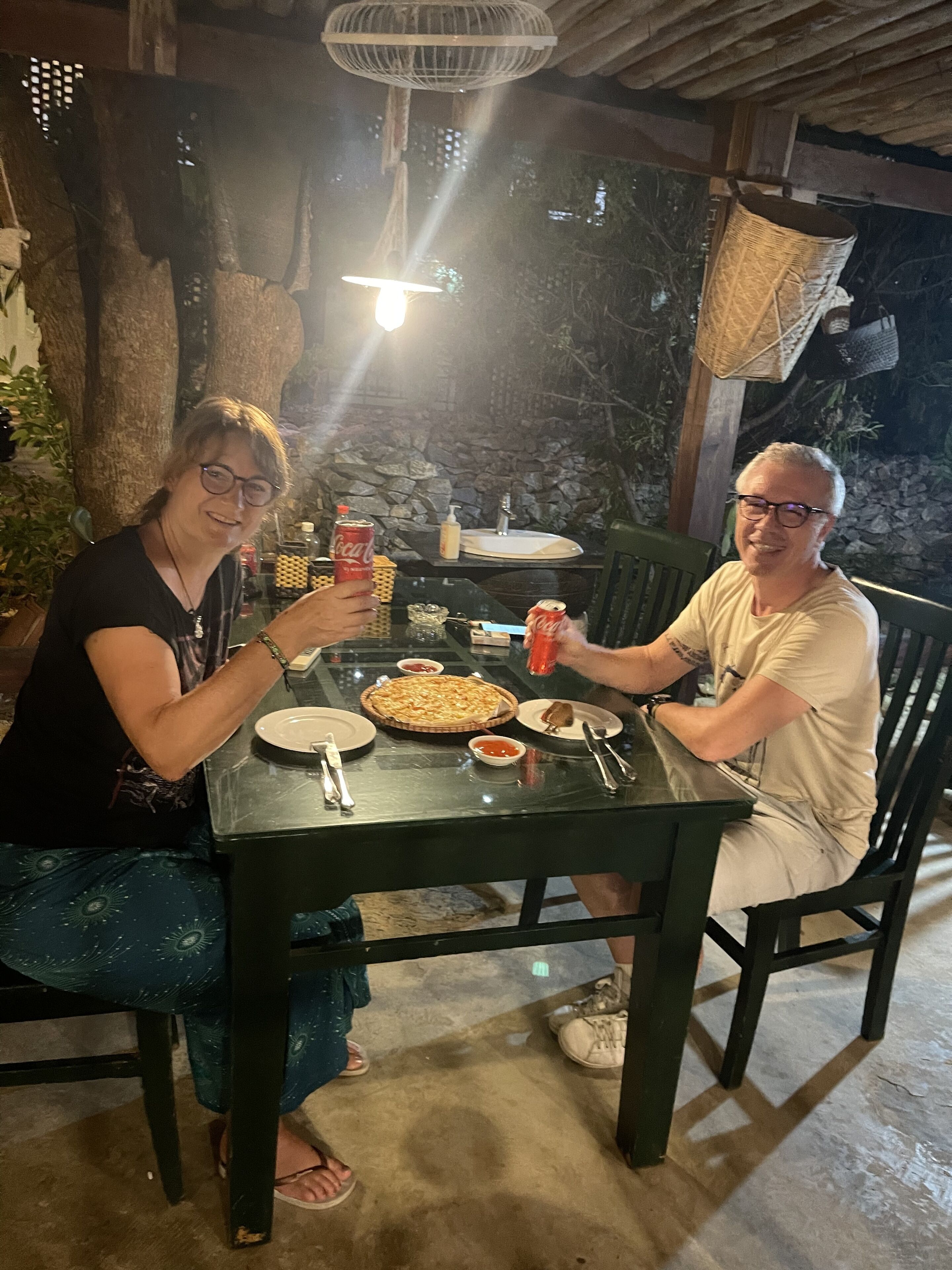 Deux personnes sont assises à une table à l’extérieur, devant une pizza, des boissons et des accompagnements.