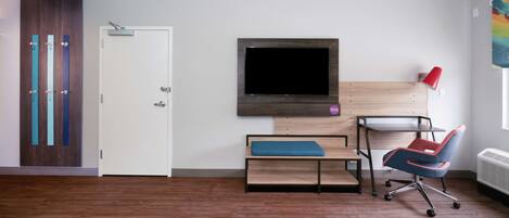 A modern room with a wooden floor, a white door, a TV mounted on a wooden panel, a desk with a chair, and a red lamp.