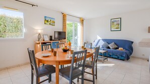 Een eethoek met een ronde tafel, stoelen en een televisie. Er is een blauwe bank en een raam met gordijnen.