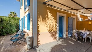 Een overdekt terras met een tafel en stoelen, een deur en een raam met blauwe luiken.
