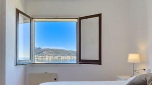 A hotel room with a bed, a nightstand, a lamp, and a large window offering a view of a coastal town. - Apartment Rhodas by Interhome (Roses)