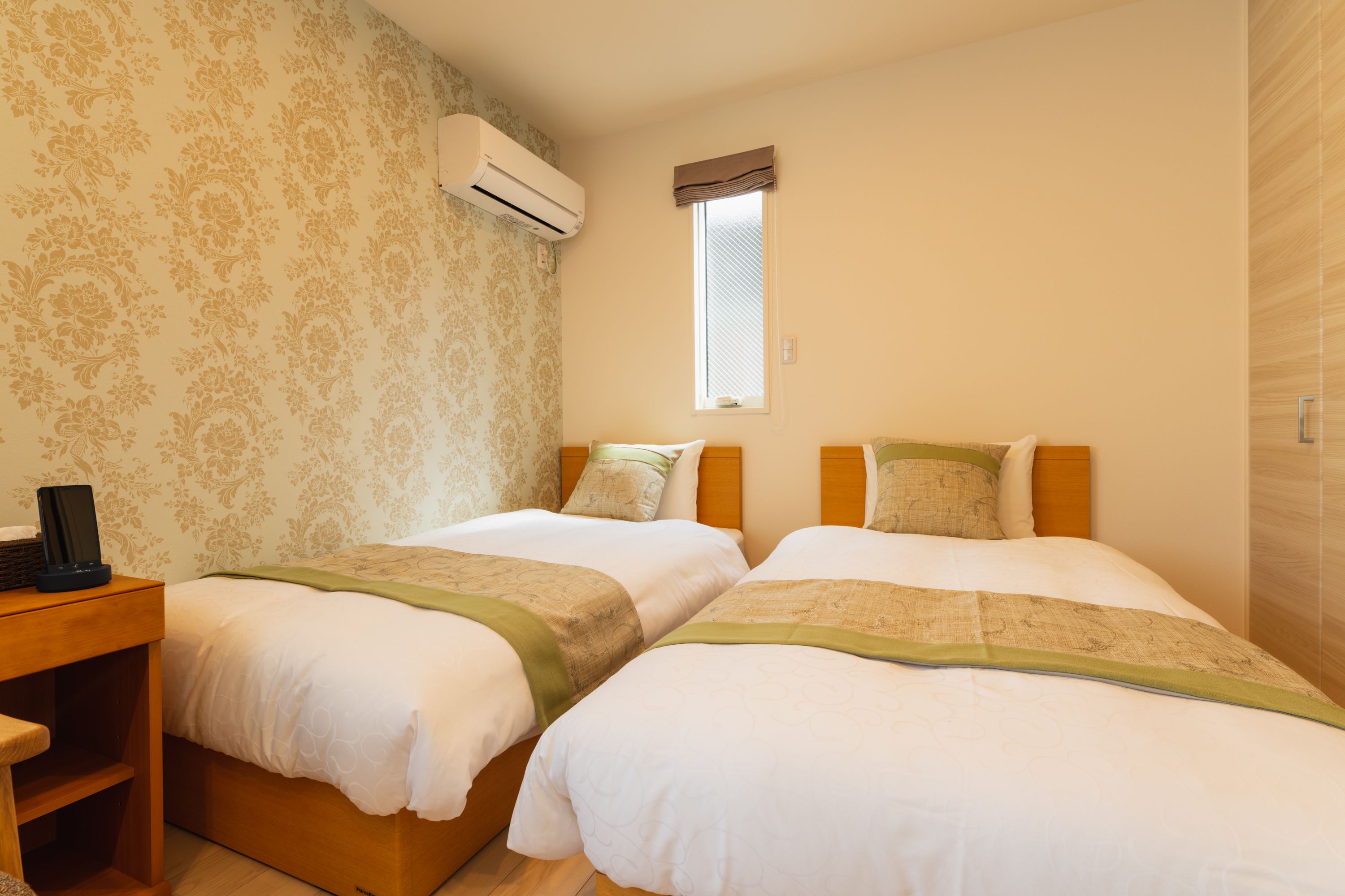 Une chambre d’hôtel avec deux lits, une table de chevet en bois, un appareil de climatisation et une fenêtre agrémentée d’un lambrequin marron.