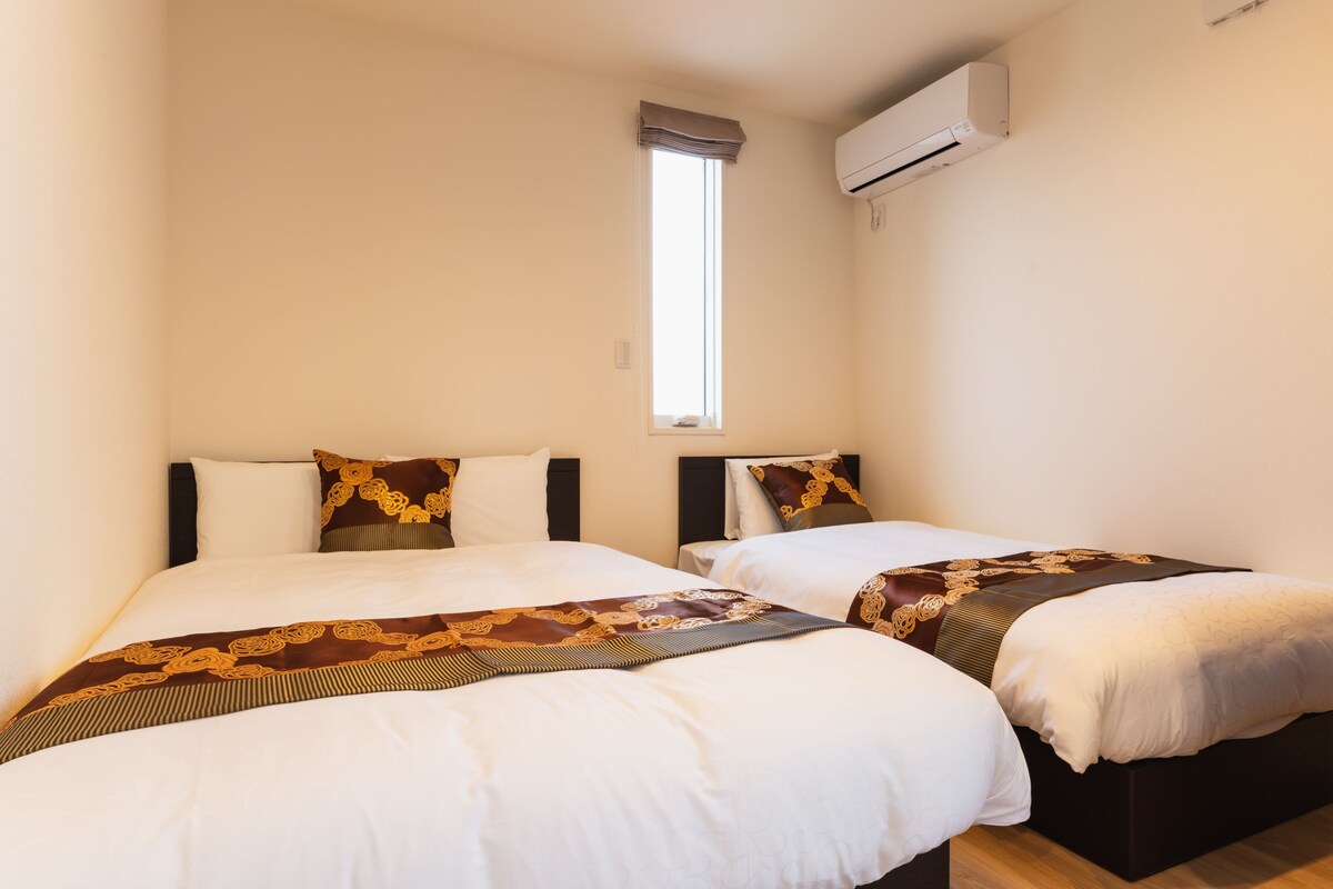 A hotel room with two beds, each with white bedding and brown decorative pillows. There is a window with a brown valance and an air conditioning unit mounted on the wall.