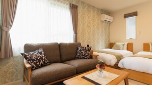 A hotel room with a sofa, two beds, a wooden coffee table, and a window with curtains.