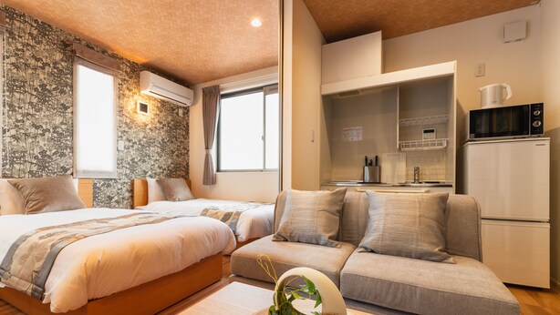 A modern bedroom with two beds, a sofa, a kitchen area, and a decorative plant.