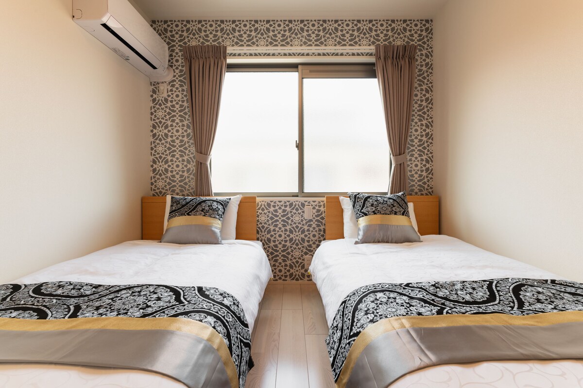 A hotel room with two beds, patterned curtains, an air conditioning unit, and a window with a view.