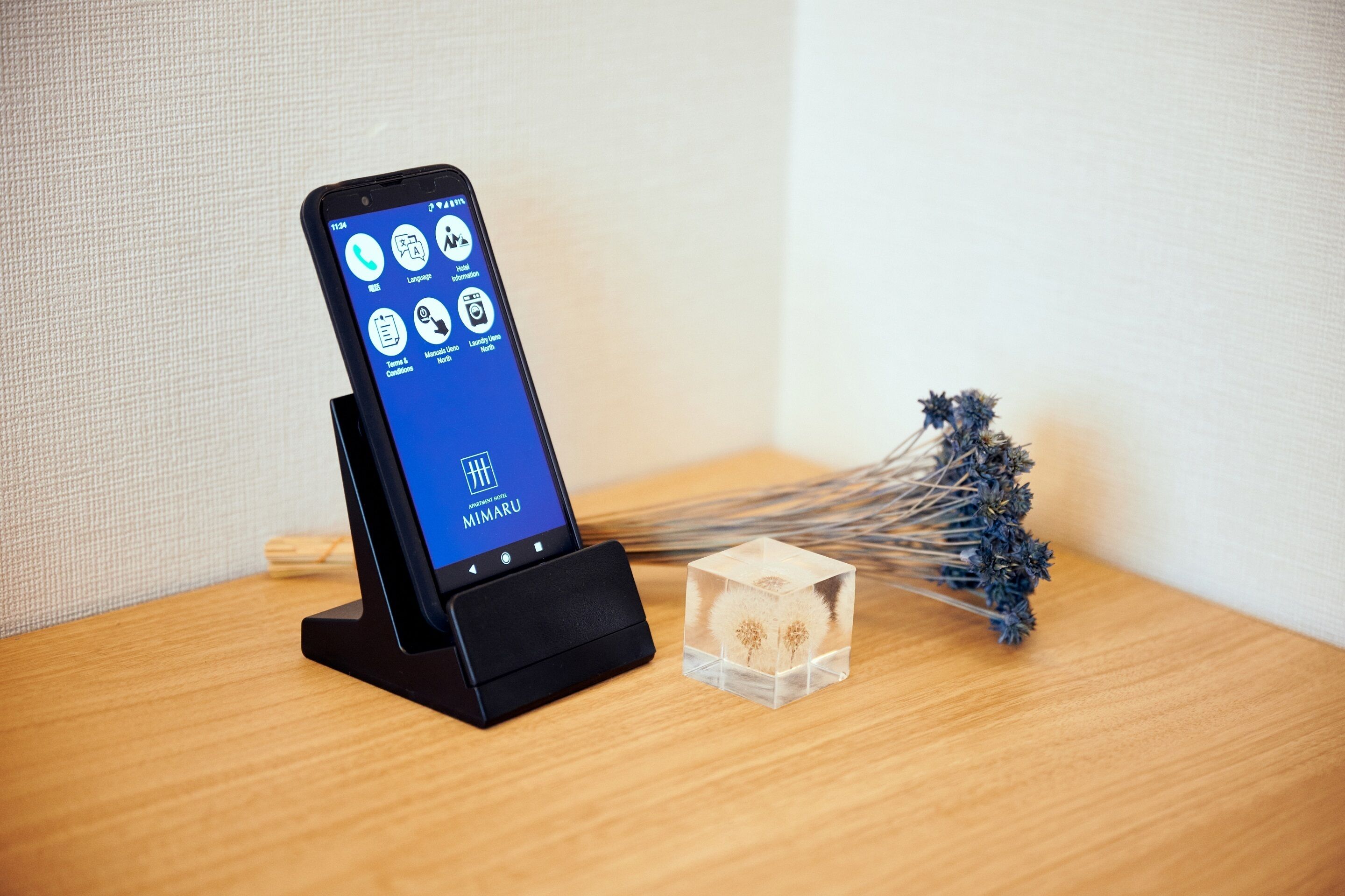 Un teléfono inteligente en una base de carga con pantalla visible, un pequeño cubo con flores secas y una superficie de madera.