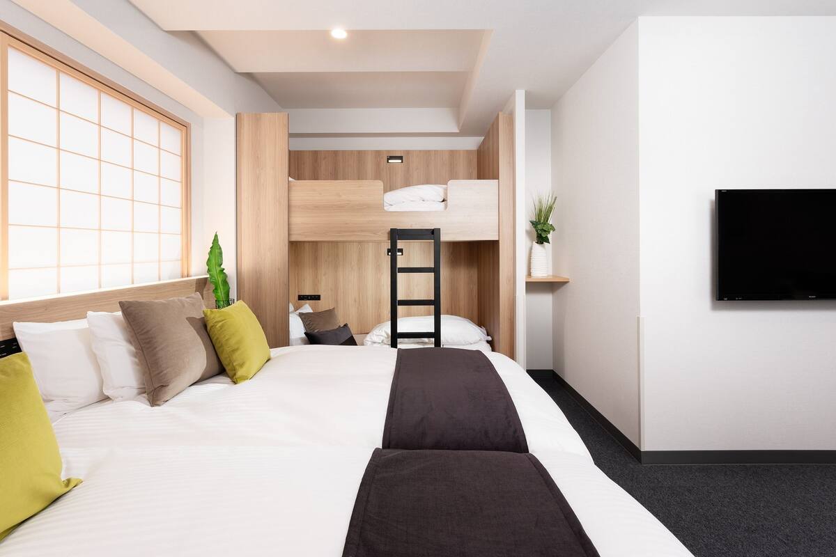 Une chambre d’hôtel moderne équipée d’un lit superposé, d’un téléviseur à écran plat fixé au mur et d’une grande fenêtre laissant entrer la lumière naturelle.