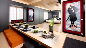 Una habitación de hotel moderna con una amplia mesa de comedor de madera, una cama con edredón blanco y obras de arte en las paredes.