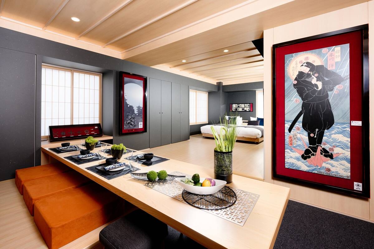 Une chambre d’hôtel moderne avec une grande table à manger en bois, un lit recouvert d’une couette blanche et des œuvres d’art accrochées aux murs.