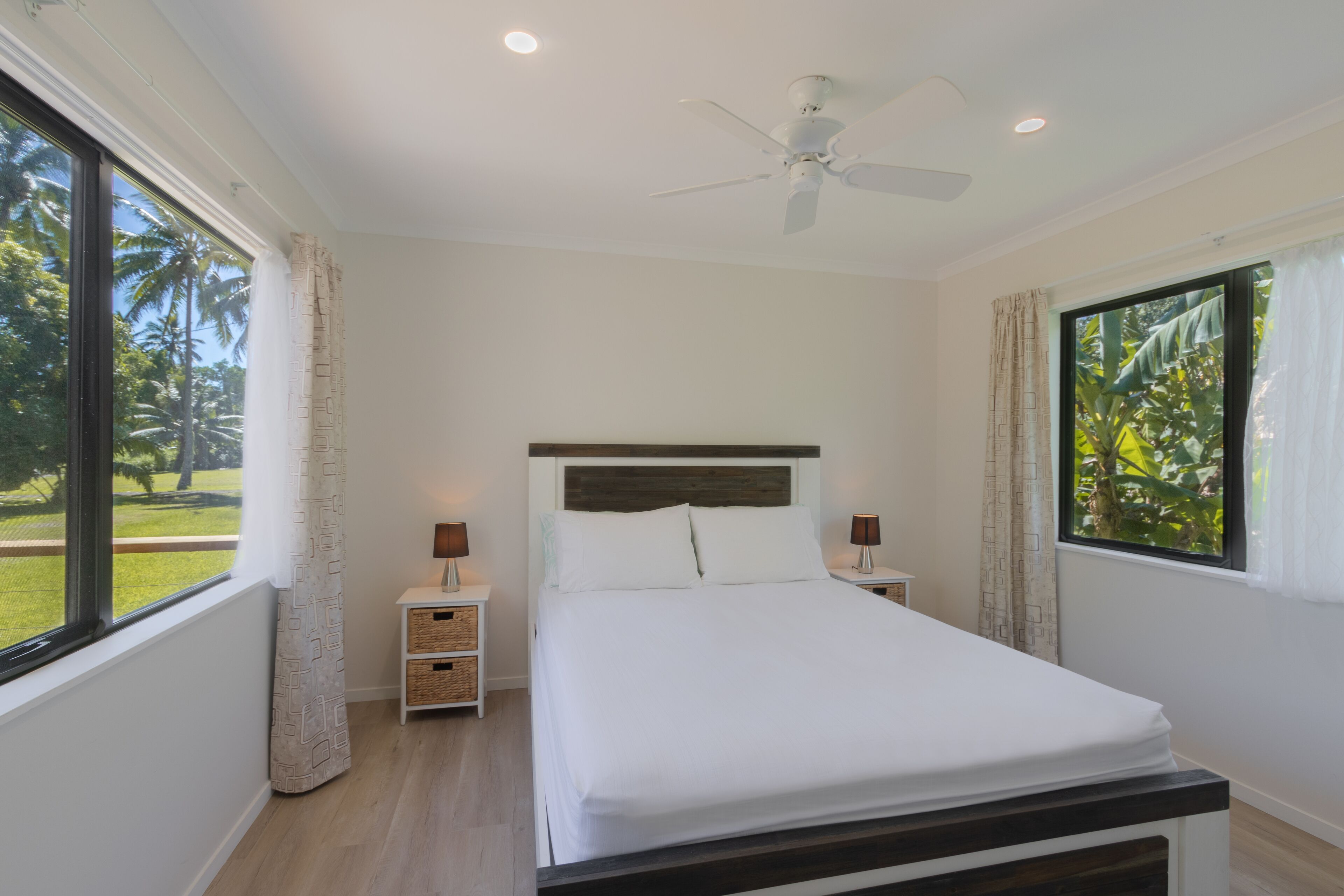 Quarto com cama, ventilador de teto, duas mesas de cabeceira e janelas amplas com cortinas.