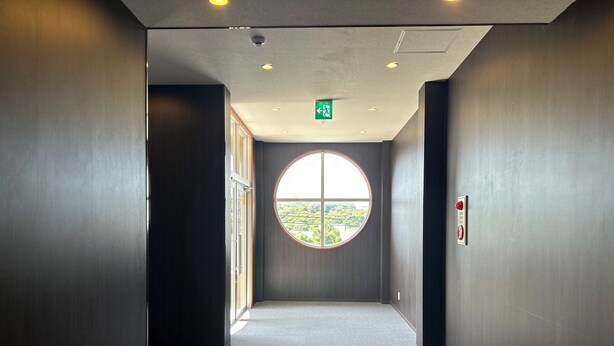 A corridor with a large circular window, a green exit sign, and a fire alarm on the wall. - Crown Hotel Okinawa Annex (Okinawa)