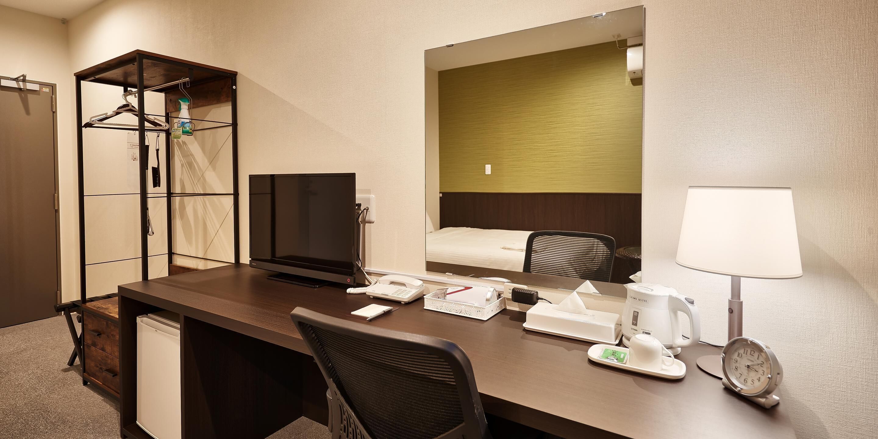 A hotel room with a desk, chair, bed, bedside table, lamp, clock, and a coat rack with hangers and a bottle.