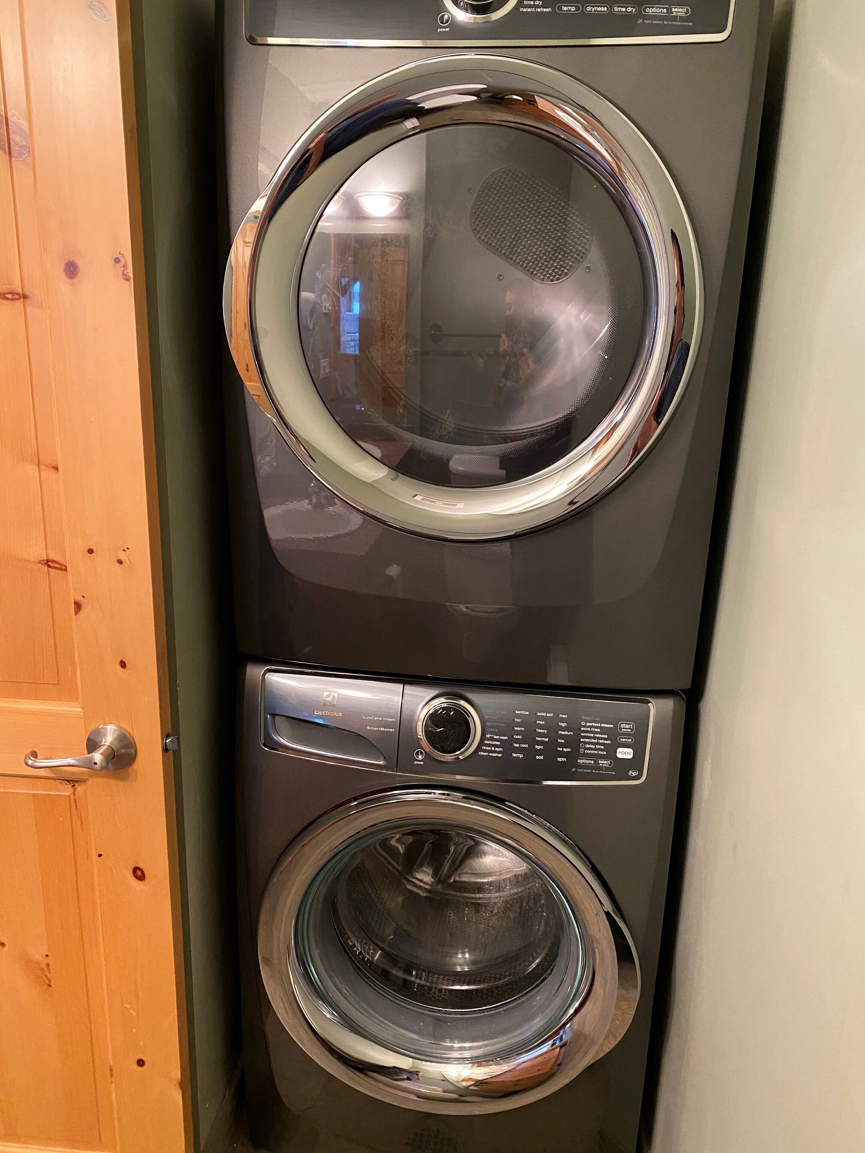 A front view of a washing machine and dryer unit with a control panel, a door handle, and a visible reflection of a person.