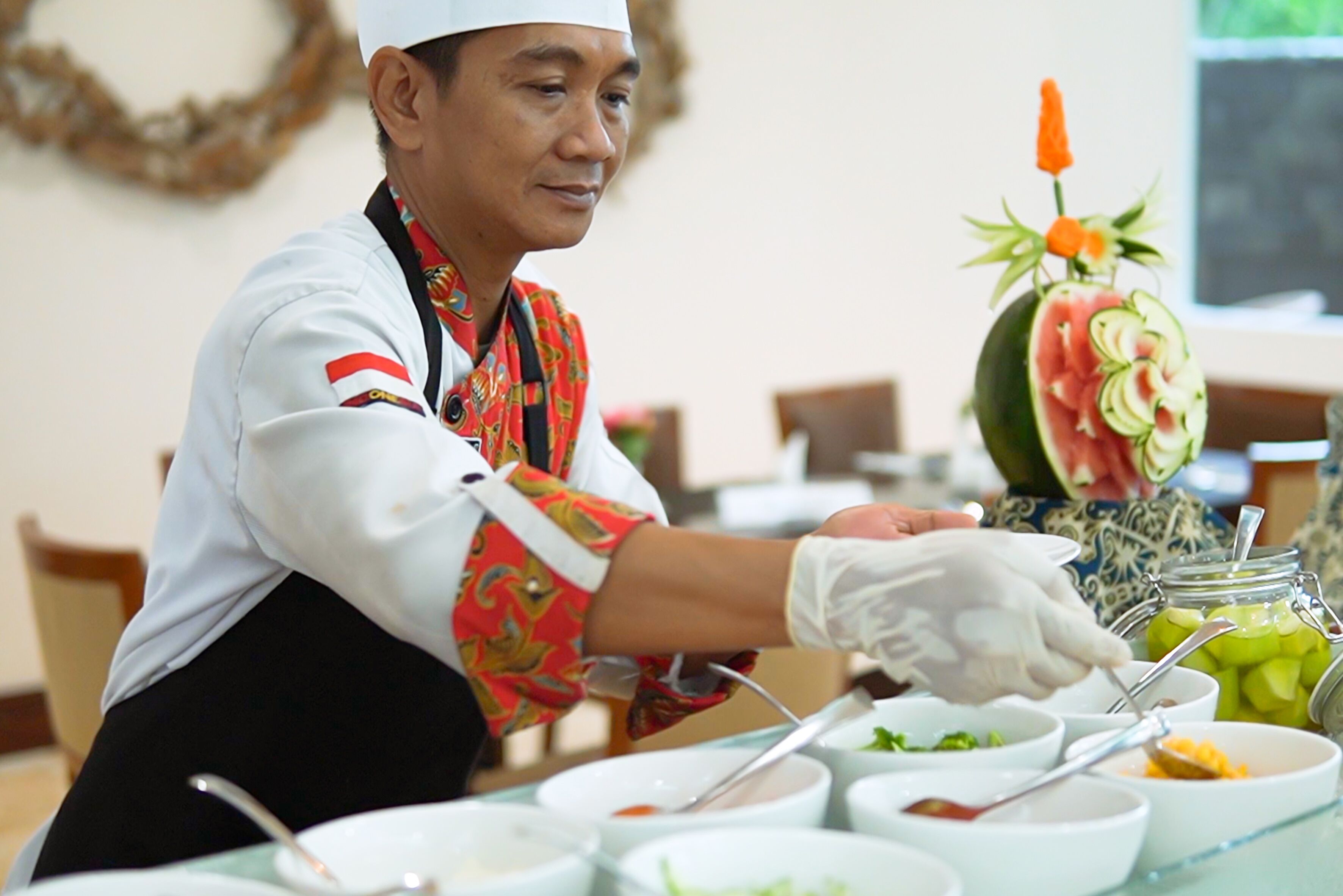 A person serving food at a buffet with various dishes and a decorative watermelon centerpiece.