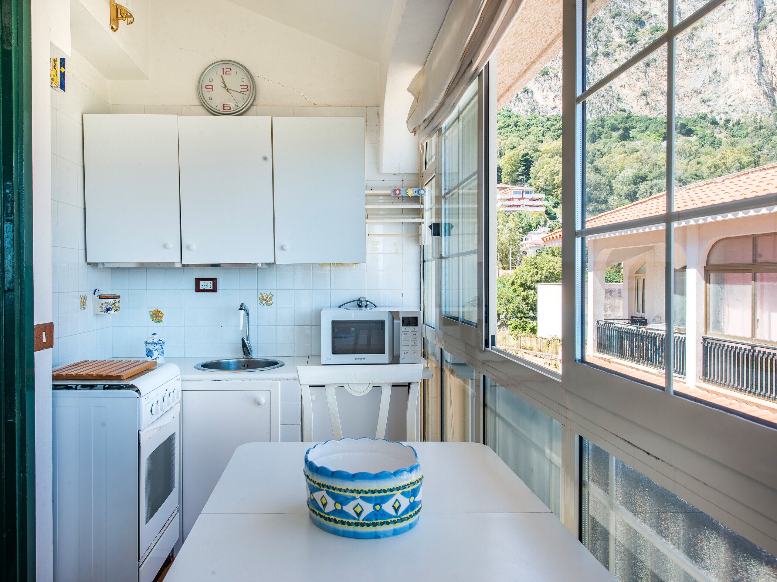 A compact kitchen with white cabinets, a microwave, and a sink. There's a clock on the wall and a window overlooking a mountainous area.