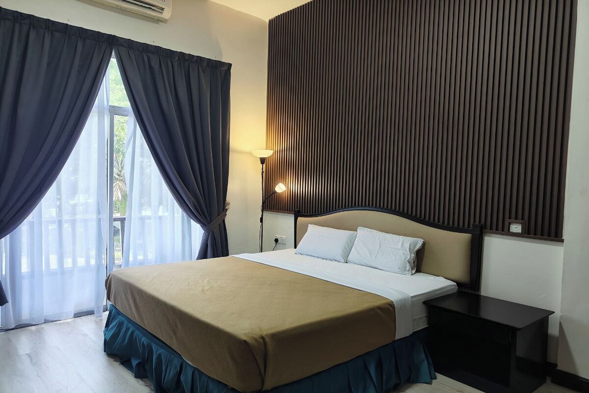 A hotel room with a bed, a nightstand, a lamp, and a view of the outdoors through a window with curtains.