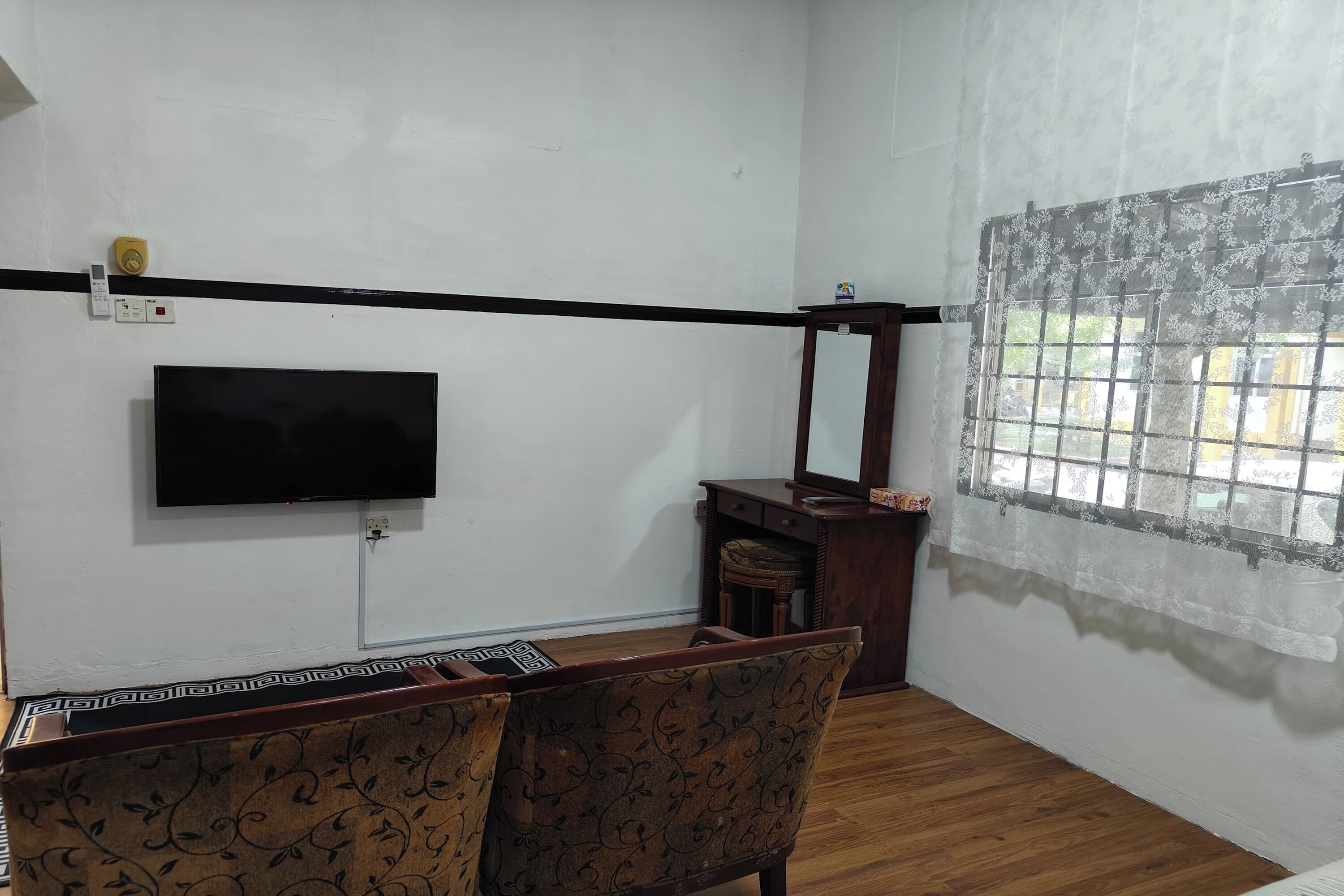 Una habitación con una televisión de pantalla plana montada en la pared, un armario de madera, una silla y una ventana con cortinas de encaje.