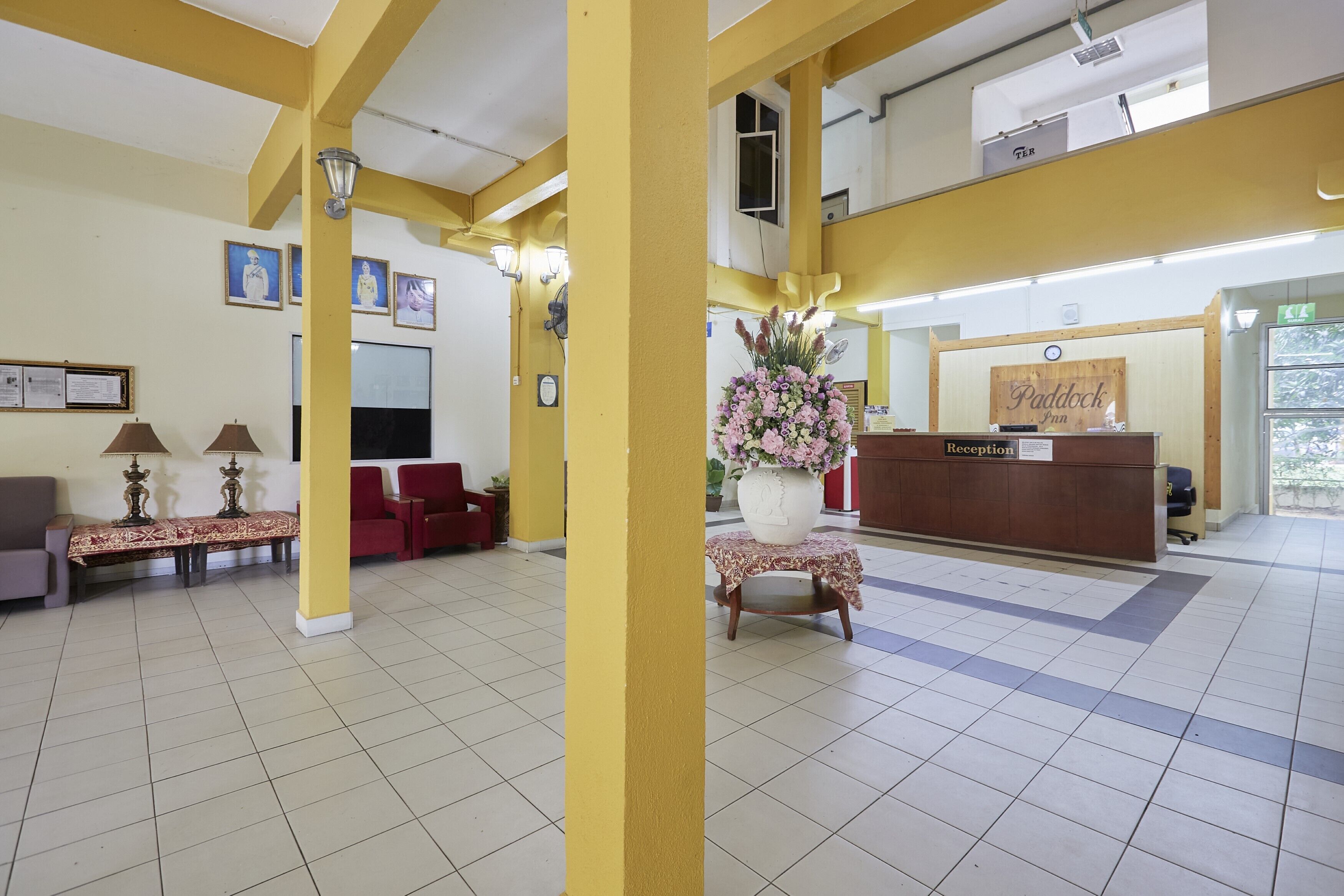 Un área de recepción con mostrador, un sofá y una silla. Hay cuadros enmarcados en la pared y un jarrón con flores sobre una mesa.
