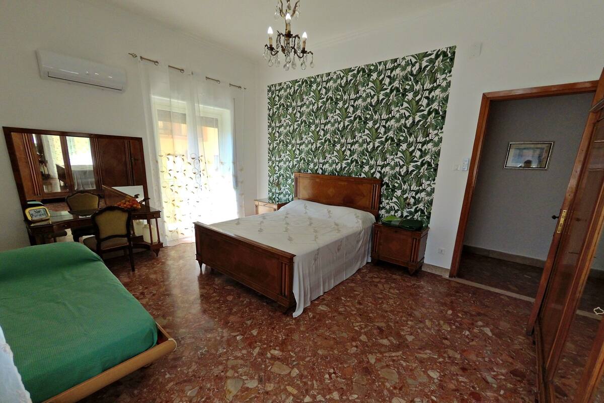 A bedroom with a bed, a chair, a desk, a mirror, and a wall with a leafy pattern.