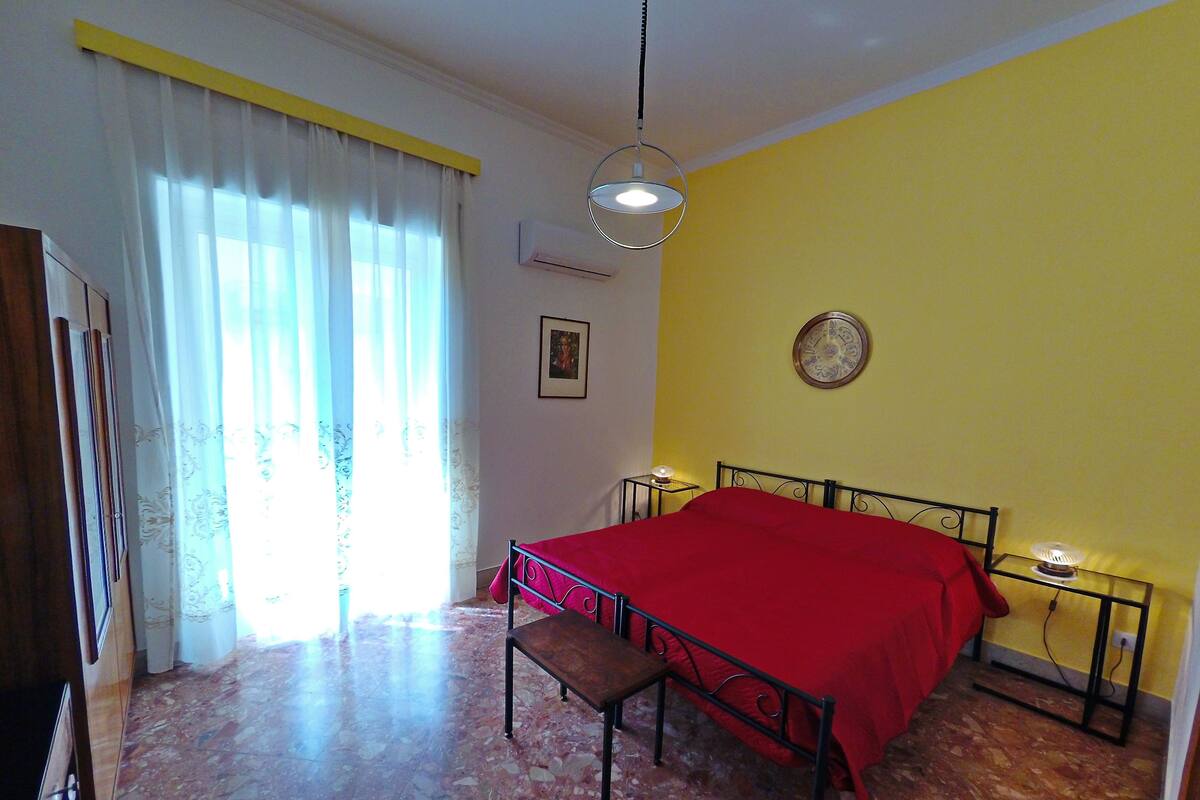 A bedroom with a bed, a bench, a bedside table with a lamp, a wall clock, and a window with curtains.