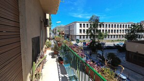 Un balcón con plantas en macetas, baranda de metal y vista a una calle con coches aparcados y edificios.
