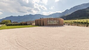 Een modern gebouw met een rond ontwerp, omgeven door een grindpad en wijngaarden, met bergen op de achtergrond.