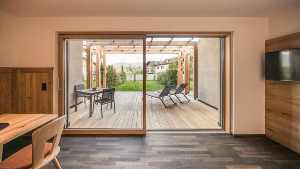 Een modern interieur met een houten eettafel en stoelen, een aan de muur bevestigde televisie, en een glazen schuifdeur naar een terras met uitzicht op een tuin en gebouwen.