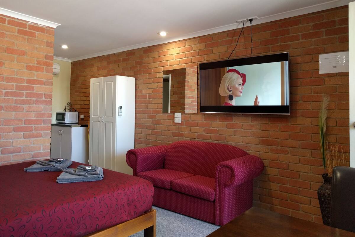 A room with a brick wall, a flat-screen TV mounted above a red sofa, a white wardrobe, and a small table with towels.