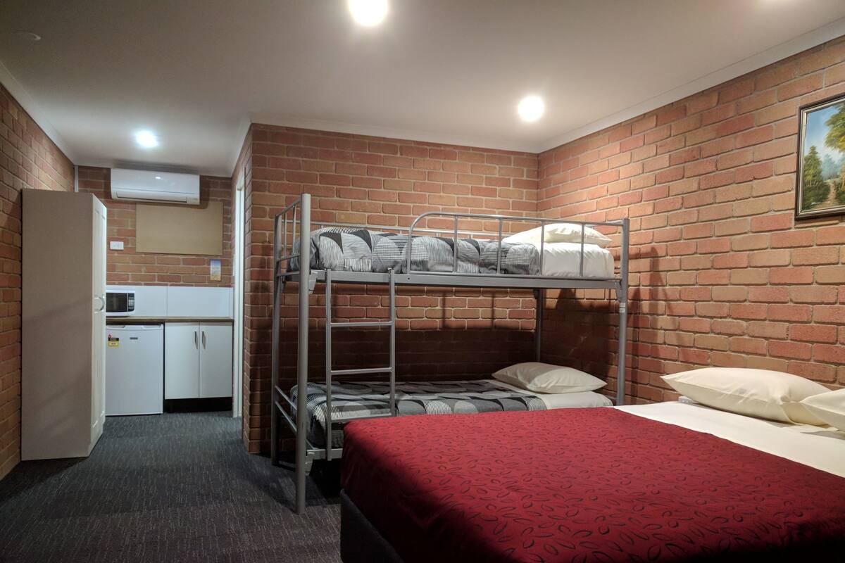 A bunk bed room with a red bedspread, a kitchenette with a microwave, and a brick wall.
