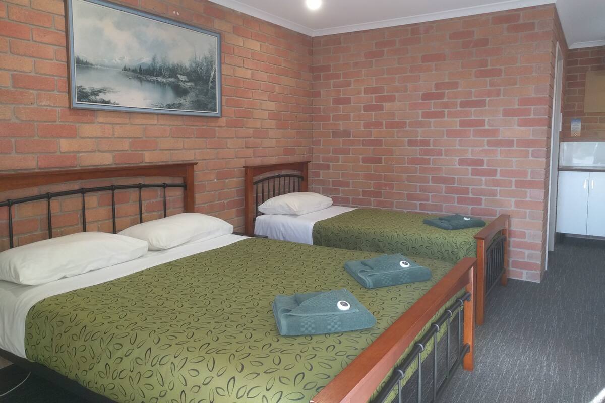 A room with two beds, a painting on the wall, and a kitchenette area.